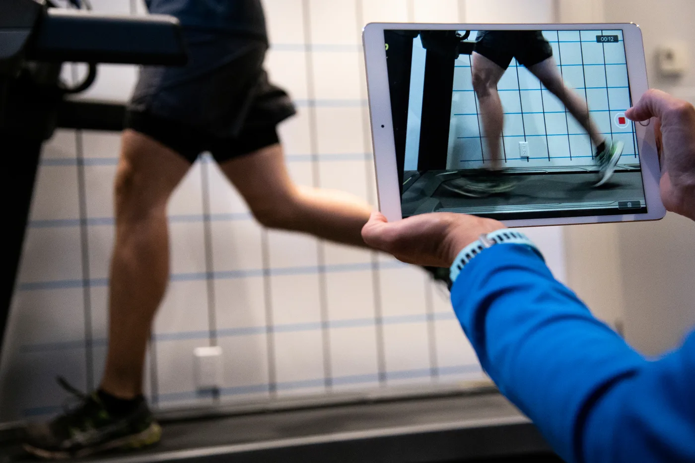 Une main tient une tablette qui filme un coureur sur un tapis roulant ; l'écran montre la grille de repères posée sur l'image