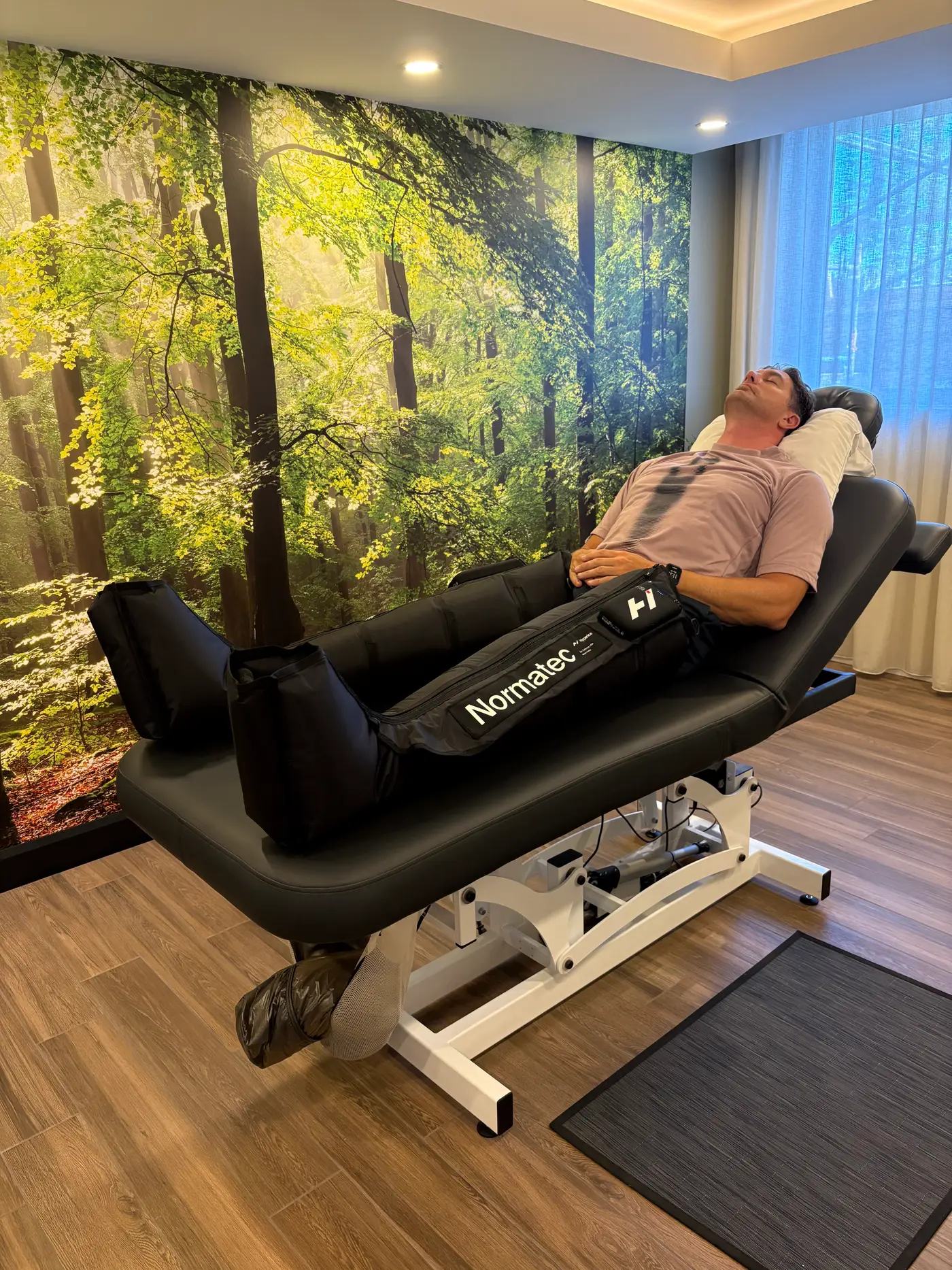 Un homme est allongé sur une table inclinée, les deux jambes dans des bottes de pressothérapie Normatec, devant une murale de sous-bois