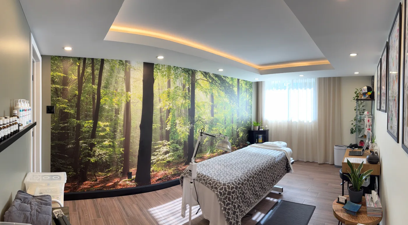 Salle de traitement : table de massage habillée d'un drap à motifs, grande murale de sous-bois sur le mur du fond, lampe sur pied, tablettes de produits à gauche