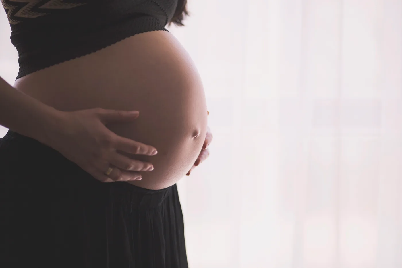 Profil du ventre d'une femme enceinte, les deux mains posées dessus, devant un rideau clair