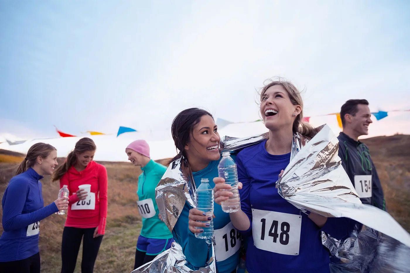 Deux femmes portant des dossards sourient après une course, enroulées dans des couvertures de survie et une bouteille d'eau à la main ; d'autres coureurs sont derrière