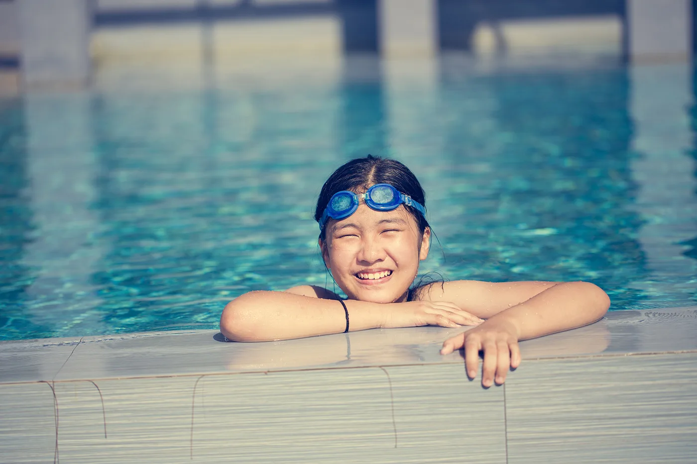 Une jeune fille avec des lunettes de natation sur le front, accoudée au rebord d'une piscine, sourit à la caméra