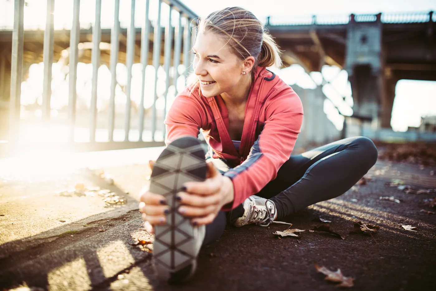 Une femme assise au sol sous un pont, jambes écartées, tient la semelle de son soulier de course en s'étirant vers l'avant