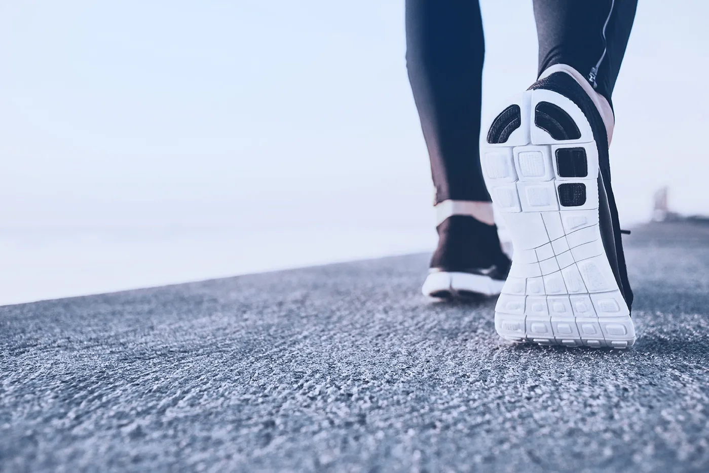 Vue rapprochée par-derrière : la semelle blanche d'un soulier de course levée au-dessus d'une route d'asphalte
