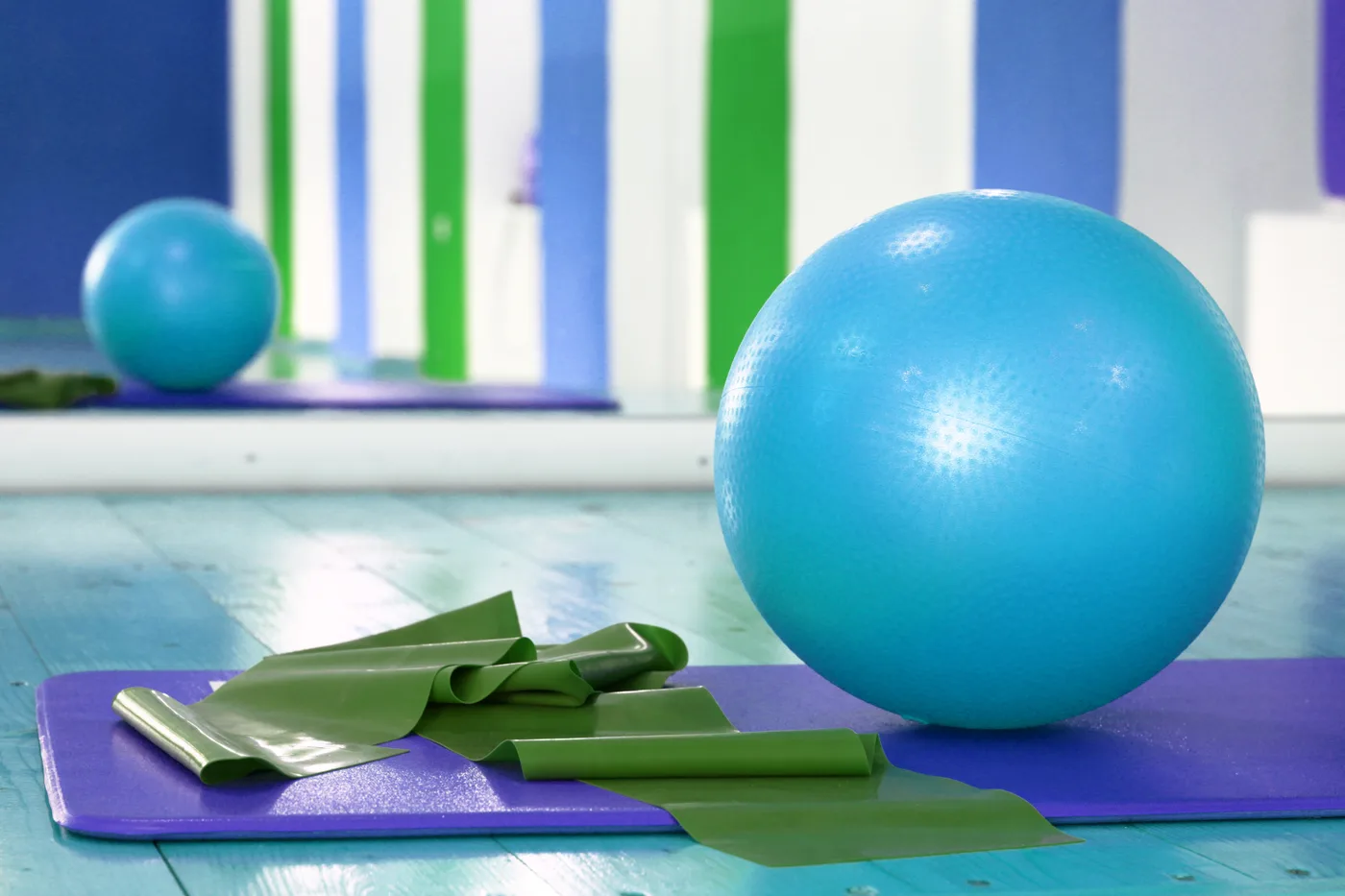 Un ballon d'exercice bleu et deux bandes élastiques vertes posés sur un tapis mauve, dans une salle aux murs bleus et verts