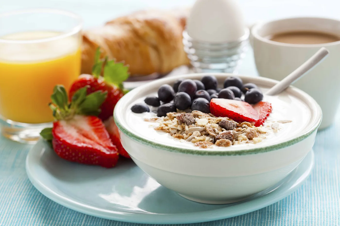 Un bol de yogourt avec céréales, bleuets et fraises, un verre de jus d'orange, un croissant, un œuf à la coque et une tasse de café