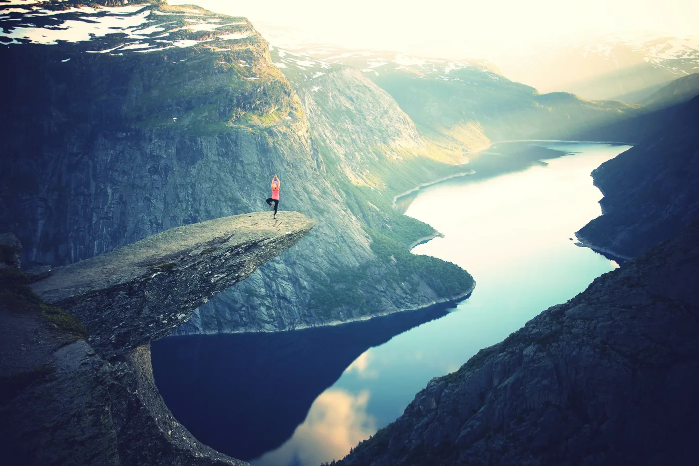 Une personne en haut rose, debout sur une pointe rocheuse en surplomb, un pied levé et les bras au-dessus de la tête ; un fjord entouré de montagnes en contrebas