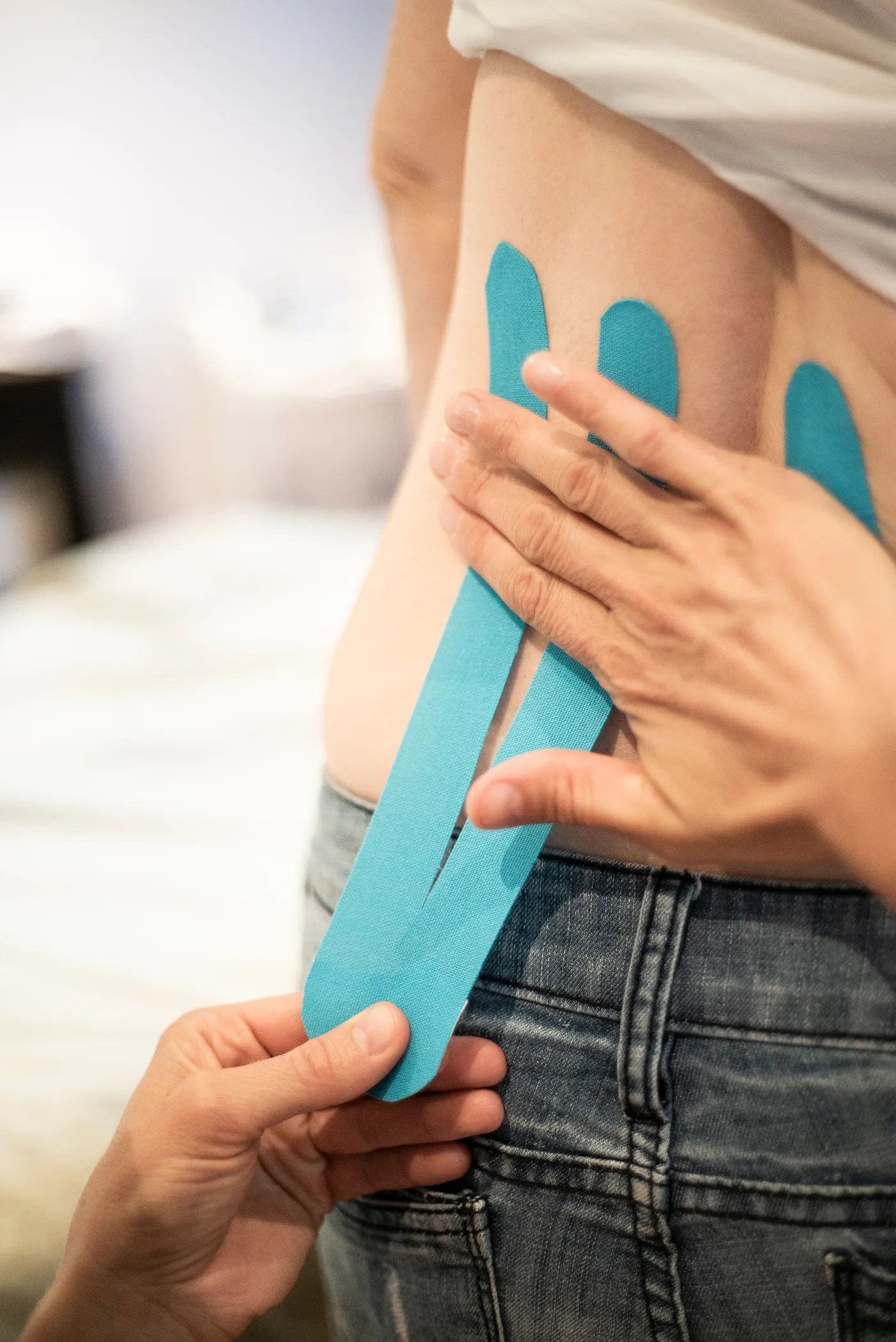Deux mains posent une bande de taping bleue sur le flanc d'une personne debout, au-dessus de la taille d'un jeans ; trois autres bandes sont déjà en place